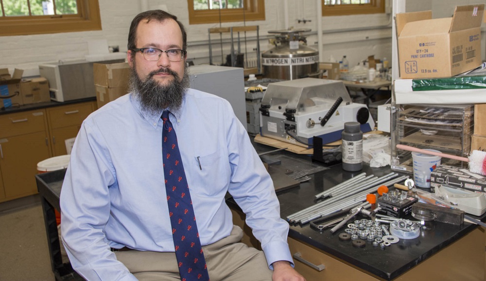 Aaron Sakulich is sitting at a lab table with a pile of small metal pieces and rods next to him. He's got dark hair, a beard, and glasses, and is wearing khaki pants, a light blue button down shirt, and a dark blue tie with red lobsters.