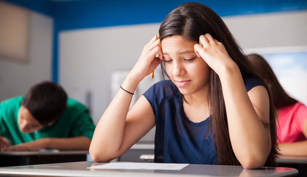 A female student studies a test on her desk. She looks concerned, and is pressing her forehead against her fists.