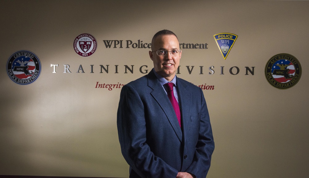 Steve Marsh stands in front of a tan wall with "WPI Police Department Training Division" written on it. He's wearing glasses and a dark blue suit and red tie, and is smiling.