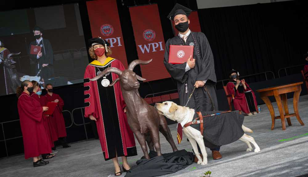 A student and their service dog stand next to President Leshin to accept their diploma.