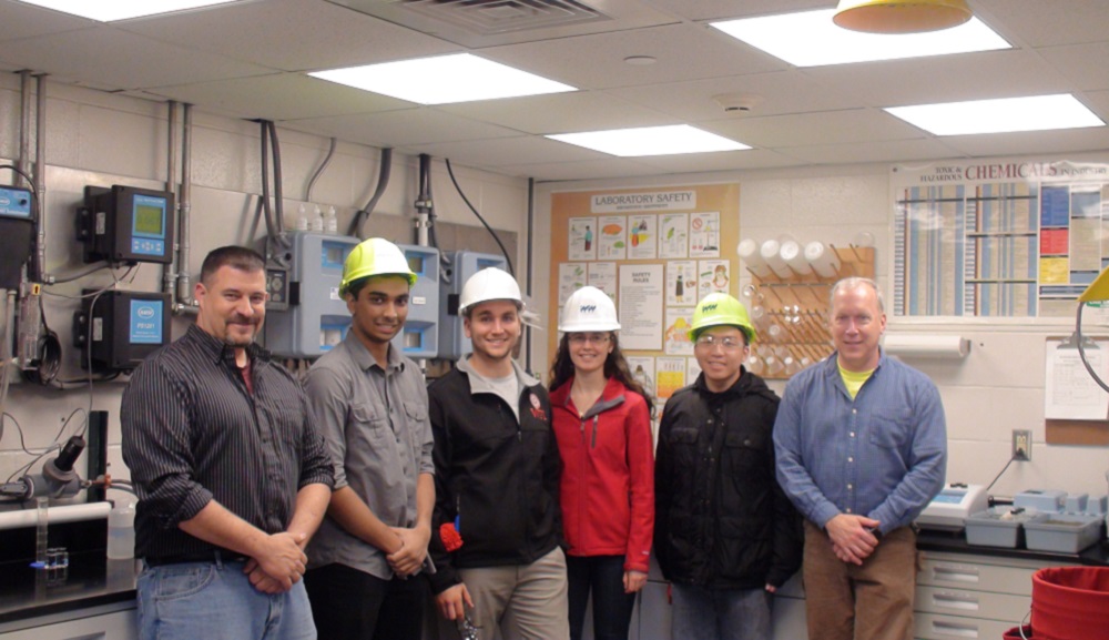 Four WPI students wearing hard hats pose with two project center sponsors in a lab area.