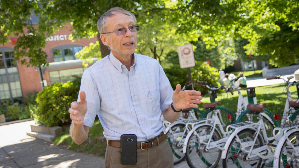 John Orr stands in front of green and white bicycles and trees that are out of focus. He's got gray hair and is wearing glasses, dark khaki pants, and a light blue checkered button-down shirt. A black cell phone case is clipped to his belt.