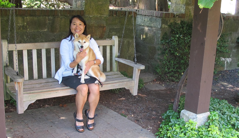 Kathy Chen sits on a wooden swing in the Higgins gardens. She is smiling and holding a dog in her lap. She's wearing a black skirt and sandals, and a light shirt and blazer. 