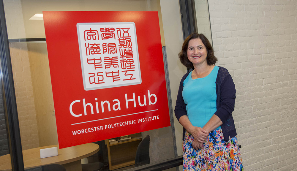Jennifer Rudolph stands next to a bright red and white China Hub sign. She is smiling, and wearing a colorful dress.