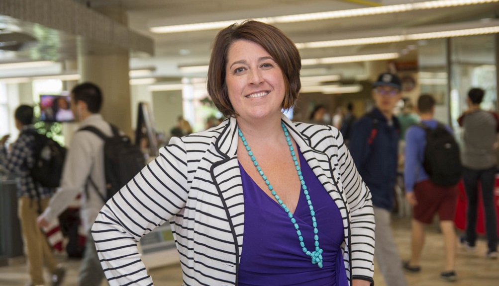 Emily Perlow stands in the Campus Center, with students walking around behind her. She is smiling and wearing a purple dress, blue necklace, and black and white striped blazer.