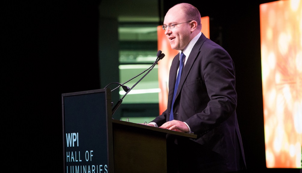 Paul Ventimiglia delivers a speech from a podium with "WPI Hall of Luminaries" emblazoned on its front. He's wearing a dark suit jacket, white shirt, and blue tie; the photo is taken from the side.