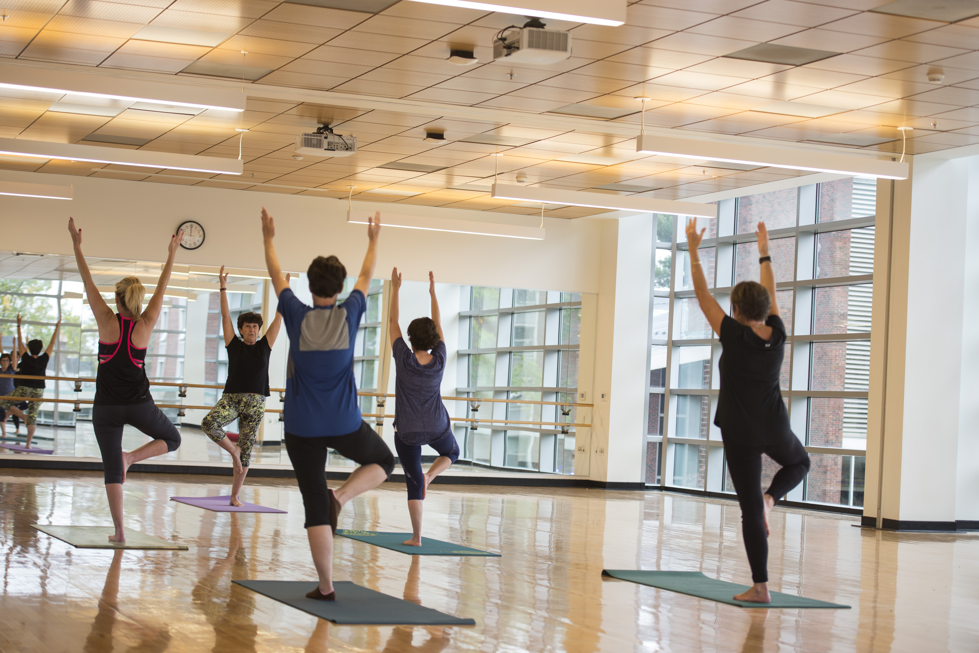 A yoga class in a light-filled studio with several women facing female yoga teacher. All have arms raised over their heads and right knee bent to the side.