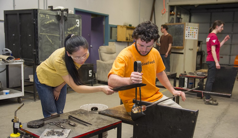 Two students participate in a glassblowing class. A male student in an orange shirt is holding a pole with a piece of glass on the end, while a female student in a yellow shirt uses a tool to hold the glass steady.