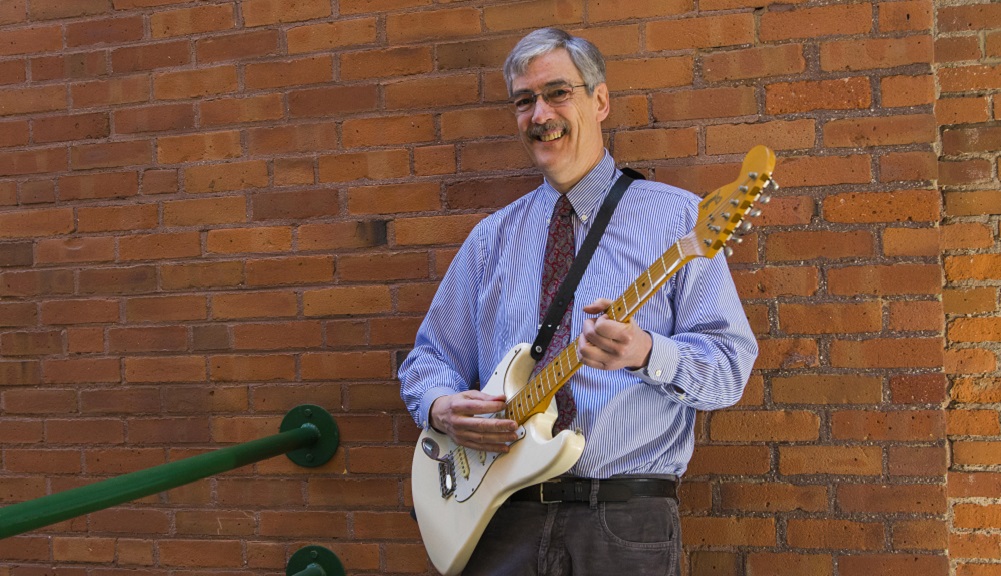 Joel Brattin stands in front of a brick wall with a guitar. He's smiling, and is wearing dark pants, and blue and white striped shirt, glasses, and red patterned tie.