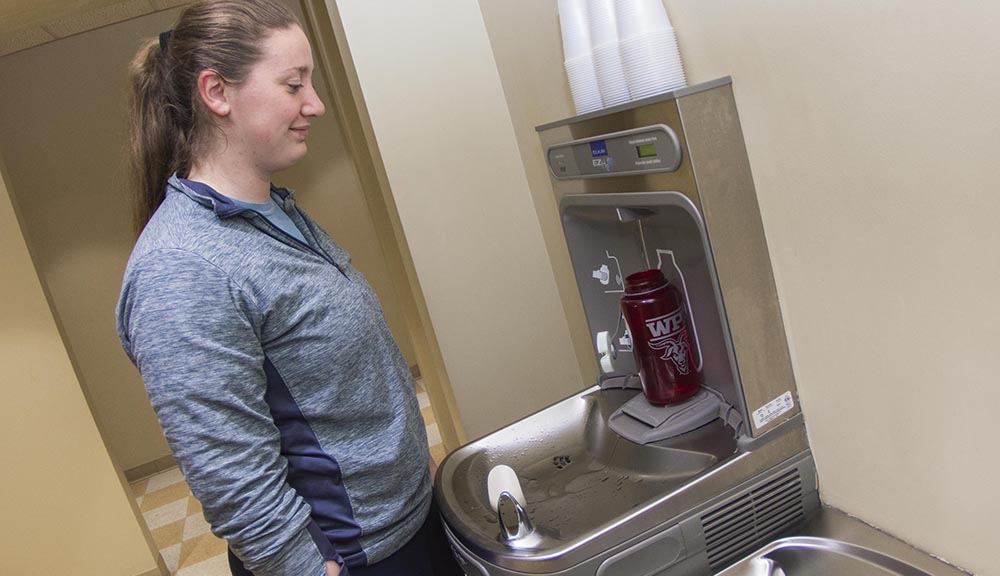 A student stands at a water fountain and prepares to refill her water bottle.