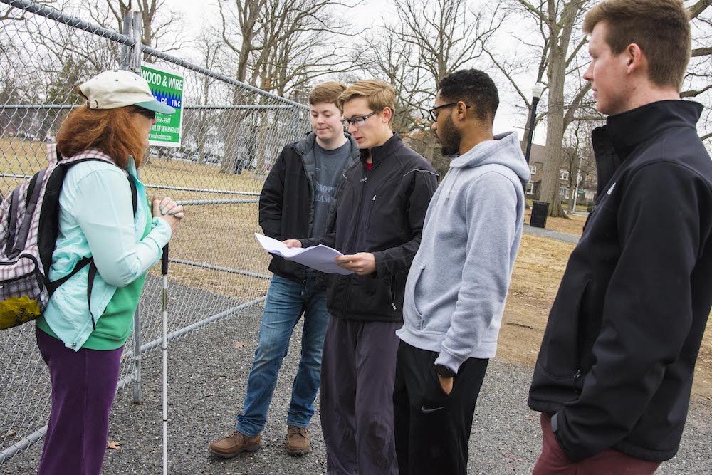 Four WPI students meet with a woman in front of a fence at Elm Park. One student is holding a map or blueprints while the rest look on.