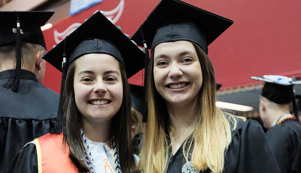 April Locke and Kelsey Leigher smile together in their Commencement caps and gowns.
