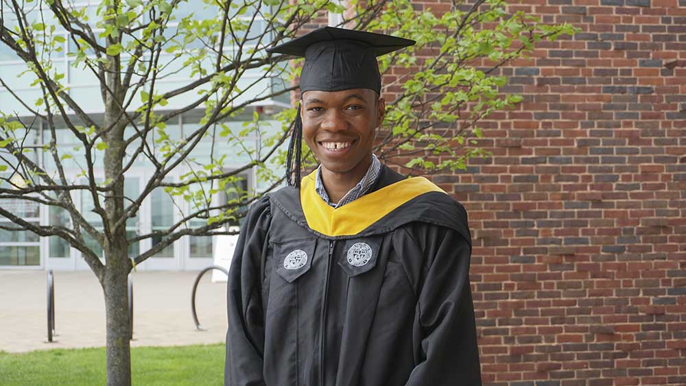 Kenechukwu Mbanisi wearing his cap and gown smiles in front of the Sports & Recreation Center.