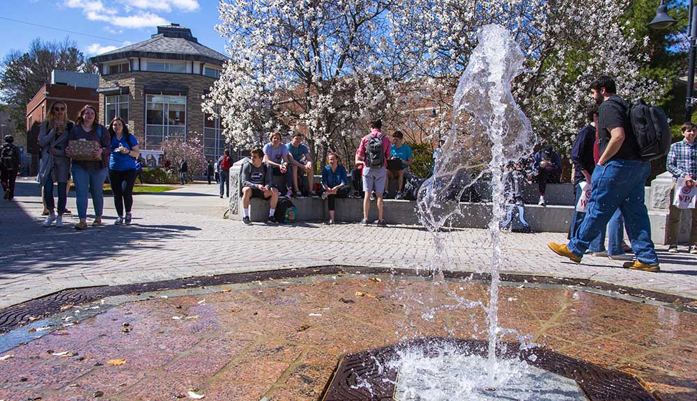 A close-up photo of the fountain the center of campus on a sunny day as students walk around and hang out together.