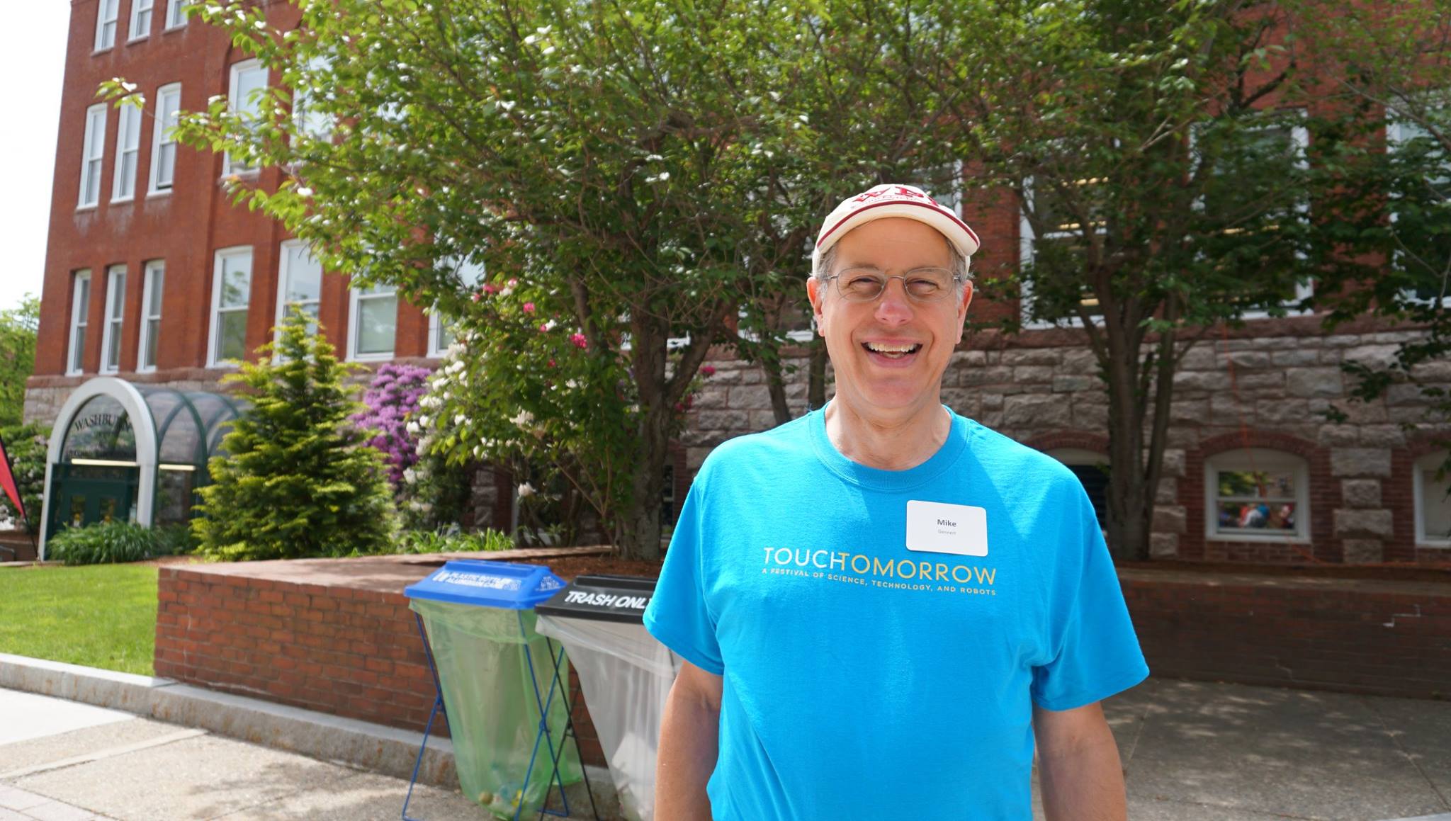 Mike Gennert stands in the center of campus. He's smiling and is wearing glasses, a WPI baseball cap, and a TouchTomorrow volunteer shirt.