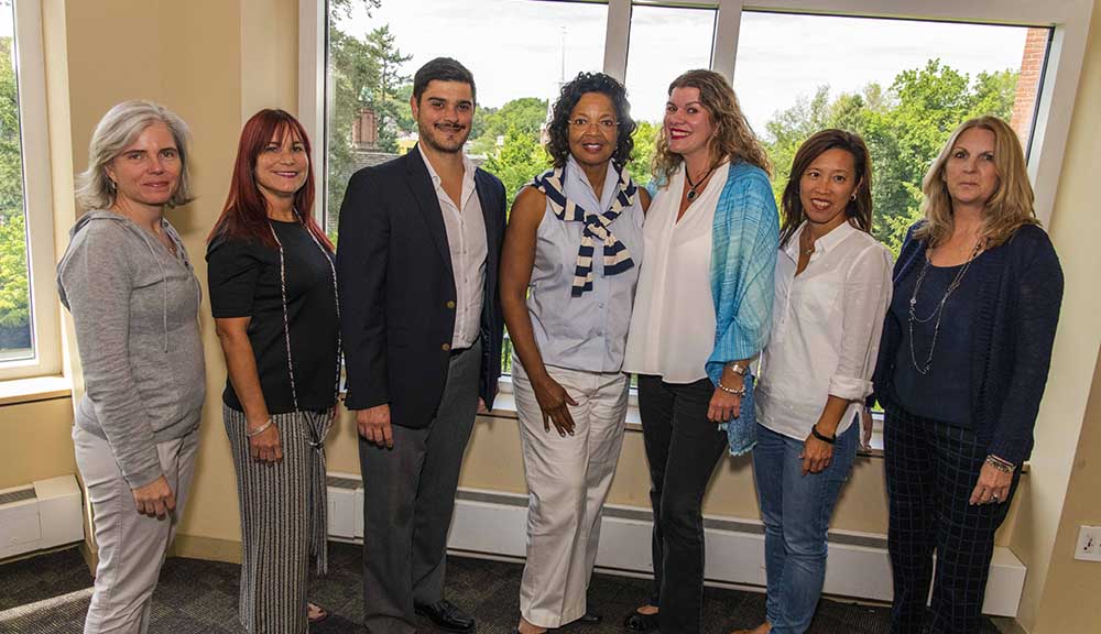 The team from Miami Dade gathers for a photo during this year's Institute on Project-Based Learning. They're wearing casual clothing and smiling, and standing in front of windows at the Rubin Campus Center.