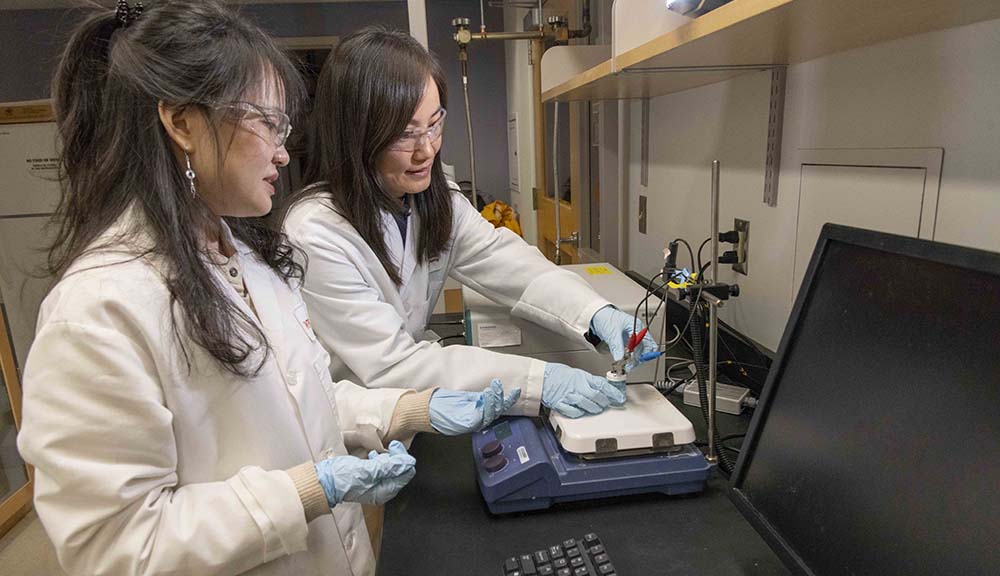 Chemical engineering professor Hong Susan Zhou, right, and PhD candidate Zhiru Zhou work together in the lab wearing white lab coats, blue gloves, and clear safety glasses.