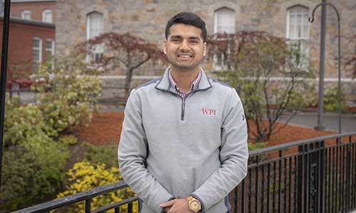 Harsh Rana stands on the Earle Bridge in front of Boynton Hall. He's smiling and is wearing a gray fleece pullover with a WPI emblem on it.