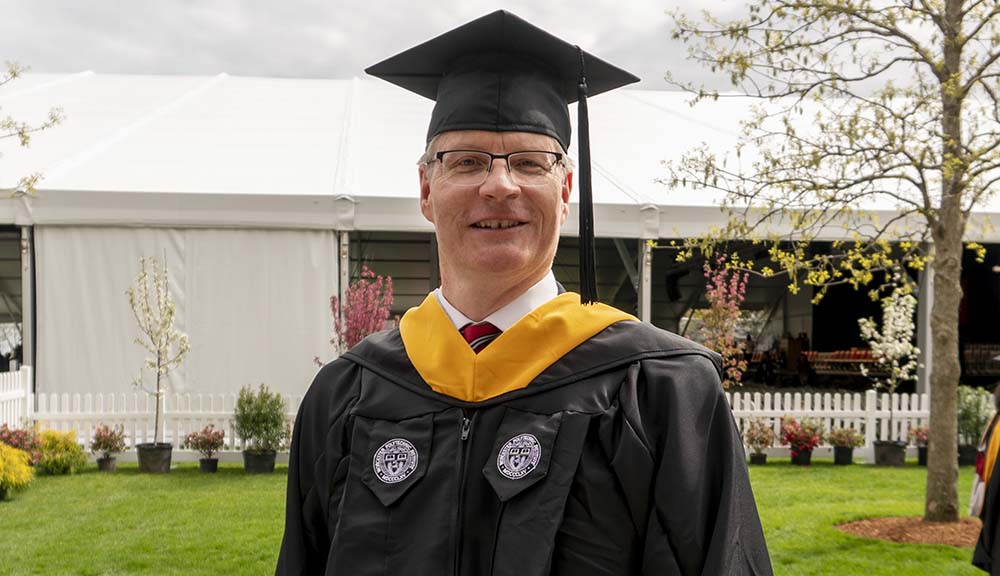 Wearing his graduation cap and gown, Andy Baron smiles in front of the Commencement tent on the quad.