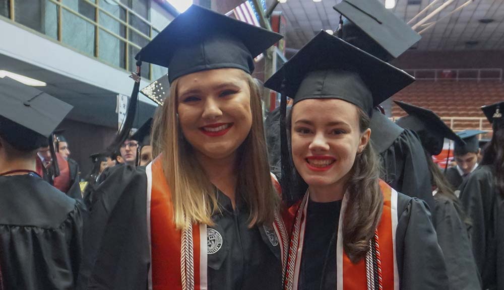 Wearing their caps and gowns, Erica Stark and Emma Travassos smile together while waiting to enter the Quad in Harrington Auditorium.