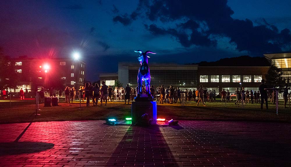 A photo taken on the Quad at night from behind the Proud Goat statue, with a variety of multicolored lights illuminating the area while students mingle.