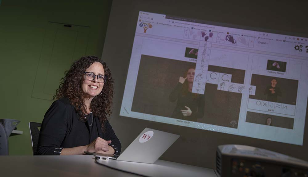 Erin Solovey sits in front of a laptop and projection screen that's showcasing someone communicating in American Sign Language.