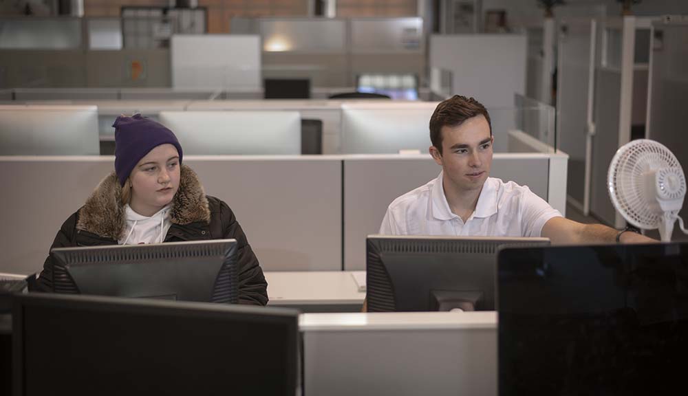 Two students (one dressed in winter clothes, the other in summer clothes) sit next to each other at computers. The student in summer clothes adjusts a fan while the student in winter clothes looks on.