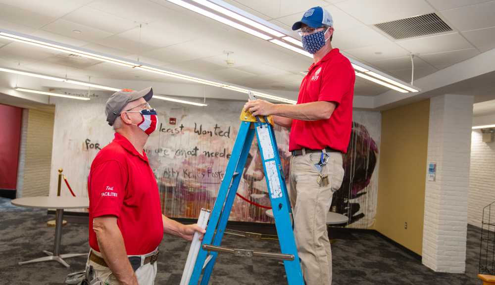 Two electricians wearing face coverings talk to each other in a WPI building. One is standing on a ladder while the other looks up at him.