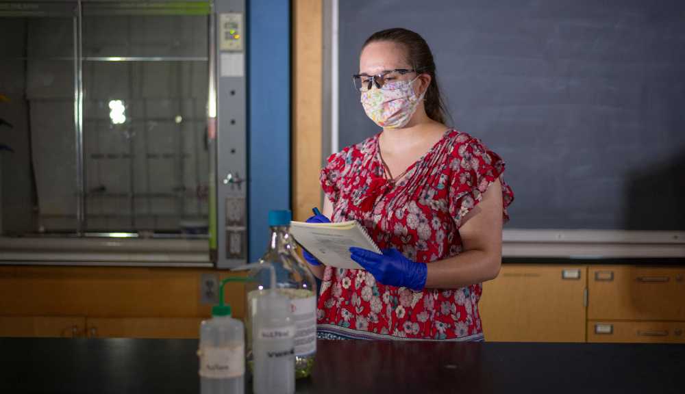 A woman wearing a face covering and gloves takes notes with a few bottles of lab chemicals on the counter in front of her.