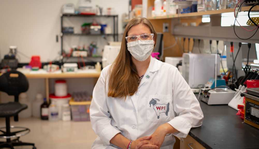Natalie Farny poses in her lab while wearing a face covering and white lab coat with Gompei on the pocket.