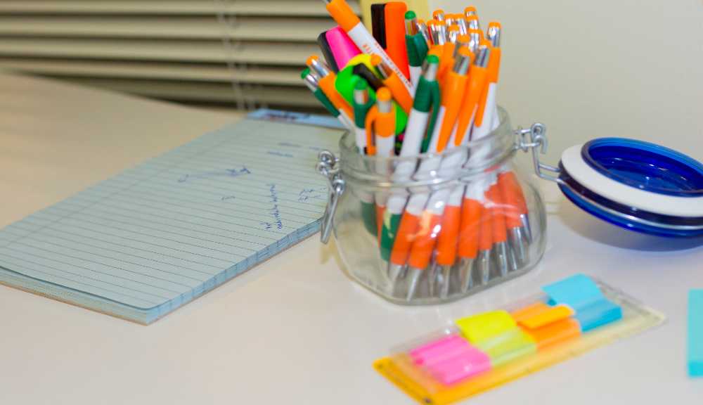 A photo of several pens in a cup on a table with sticky notes next to it.