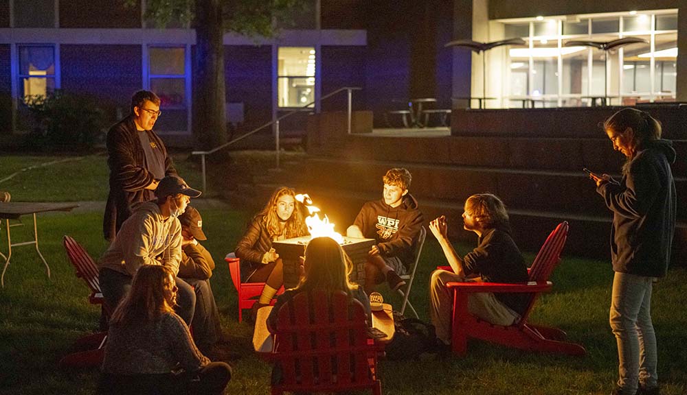 Students gather around a fire pit on campus at night.