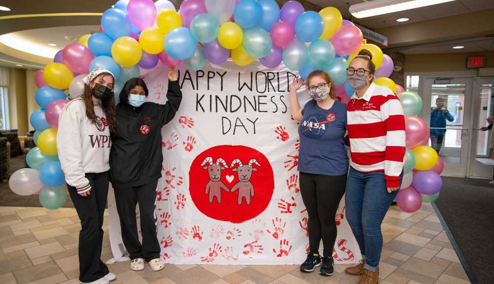 Students gather around a banner lined with balloons that reads "Happy World Kindness Day!"
