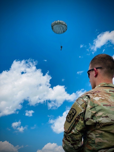 Soldier standing outside, above him is a paratrooper in a parachute