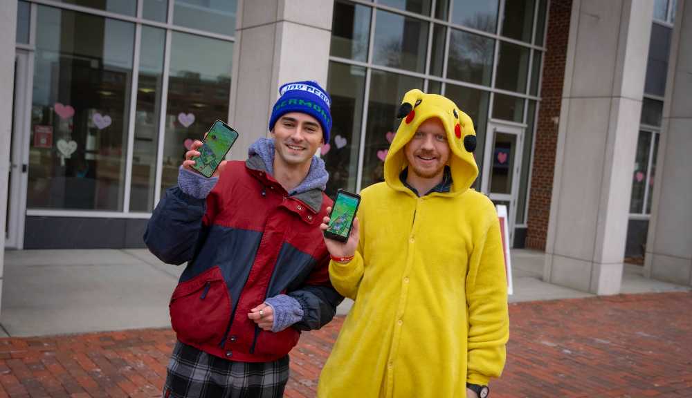 Two students, one dressed in a Pikachu onesie, hold their phones up to the camera while playing Pokemon Go.