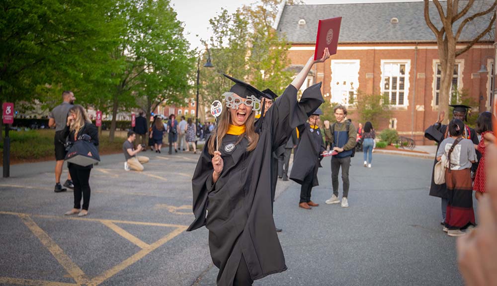 A graduate wearing "GRAD" glasses holds up her diploma in one hand and a grad sign in the other.