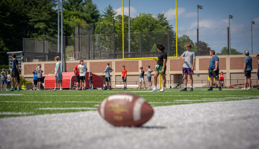 A football sits on the field while players practice in the background.