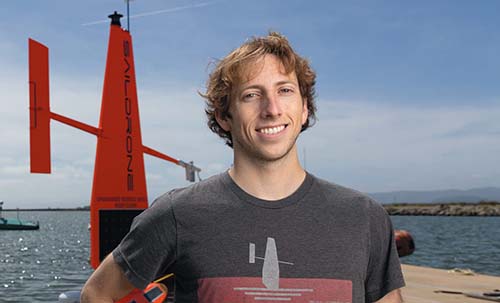 Casey Brown smiles with a blue sky and a Saildrone in the water behind him.