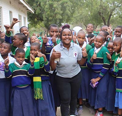Elitumaini Swai smiles and flashes a thumbs up with several young students gathered behind her. alt