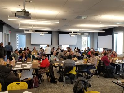 large group of faculty and staff eating lunch around tables in a large classroom before the start of the training program