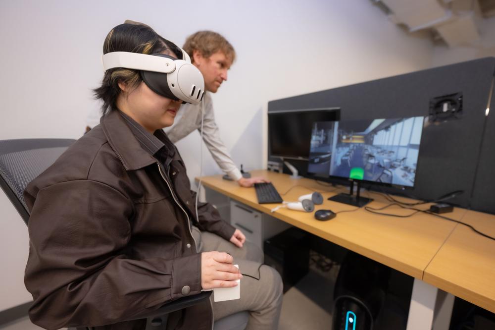 Student seated in chair, wearing virtual reality headset, operating a handheld joystick to operate the virtual reality wheelchair training system. A professor is to the left of the student, watching a computer screen with an image of what the student is seeing in her virtual reality headset, a scene of a wheelchair navigating through a restaurant.