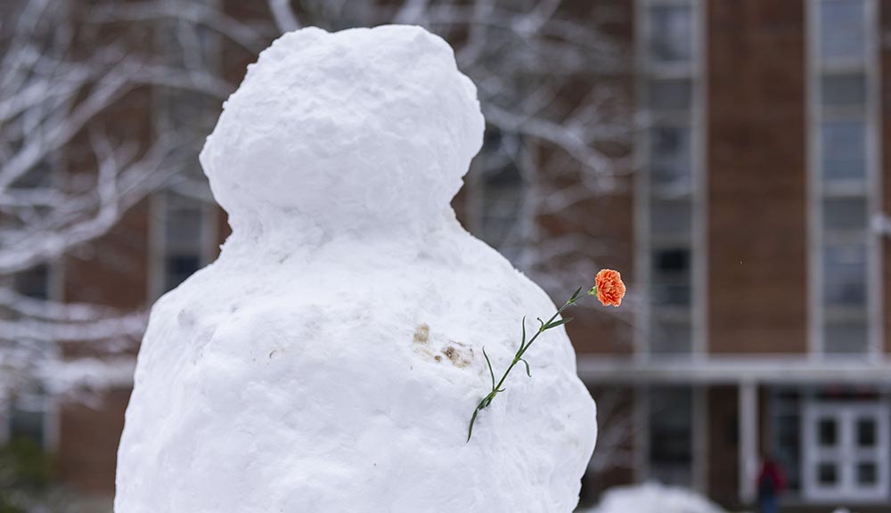 A snowman on the quad with a flower sticking out of the side.