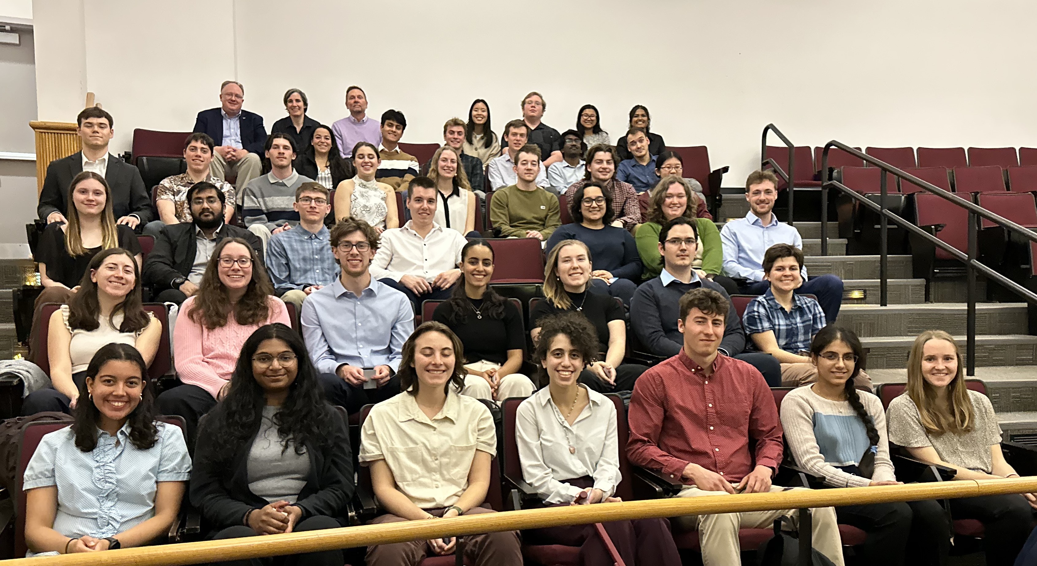 A large group of students, with a few faculty members, sits in rows of seats in an auditorium