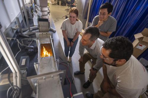 WPI researchers huddle around the outside of a wind tunnel. Inside the tunnel a plastic fuel sample is burning as part of an experiment.