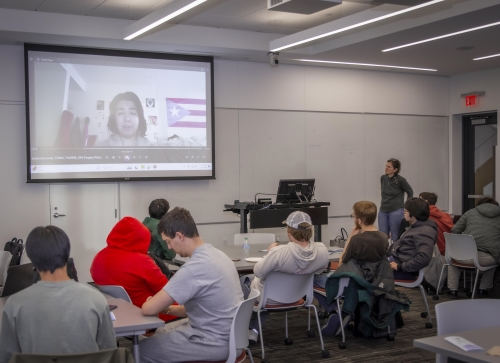 Students in classroom listening to a video presentation on a large screen from a fellow student