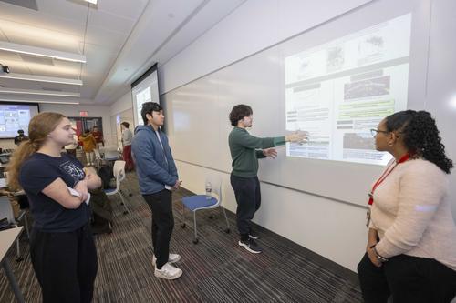 Students in a classroom present information on their projects to other students, while standing in front of a digital poster display board on the wall.