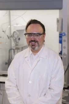 Professor Michael Timko, smiling, wearing a white lab coat and safety glasses, standing in front of lab equipment.