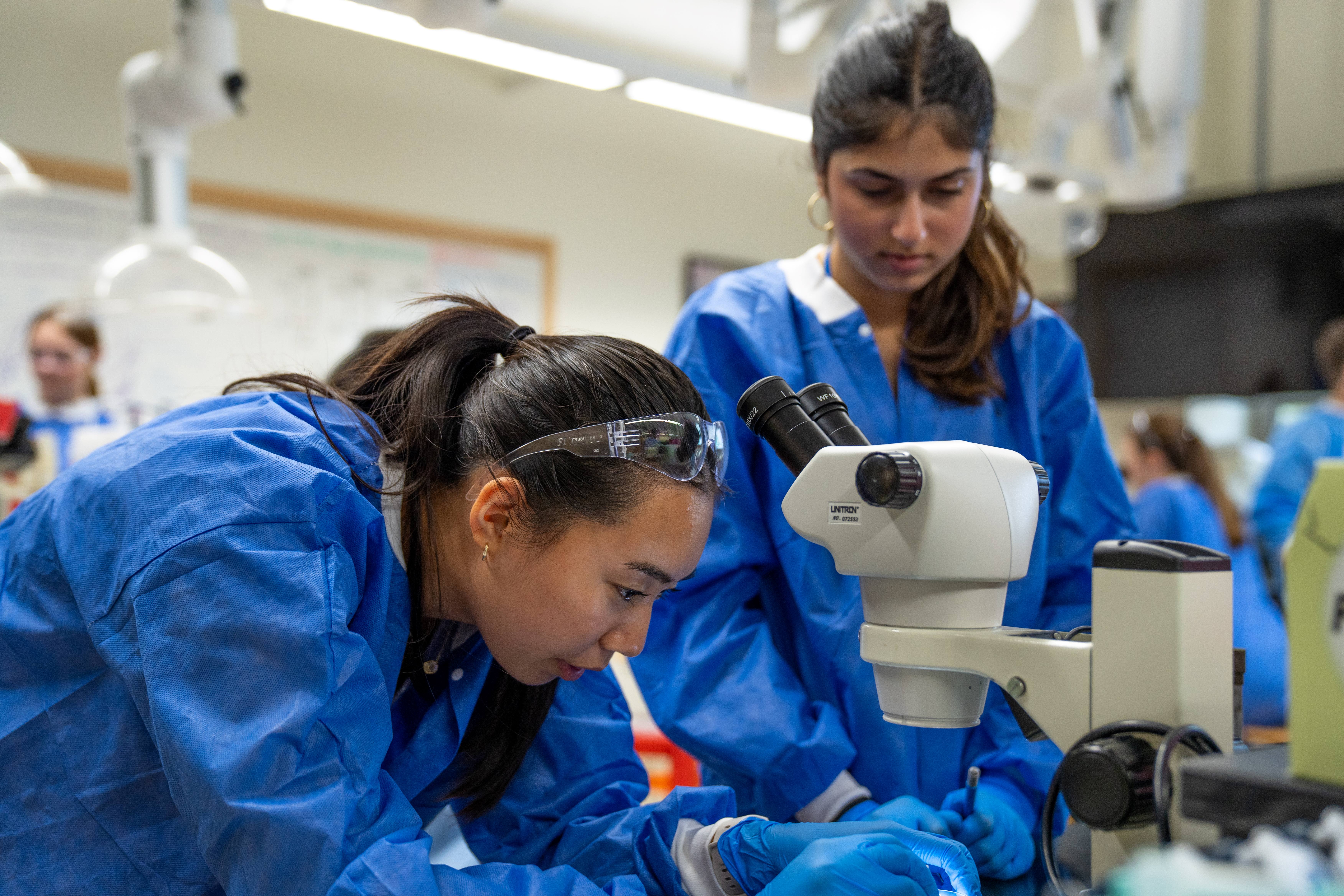 Two students wearing protective scrubs at a microscope. One is adjusting the slide under the microscope while the other student looks on.