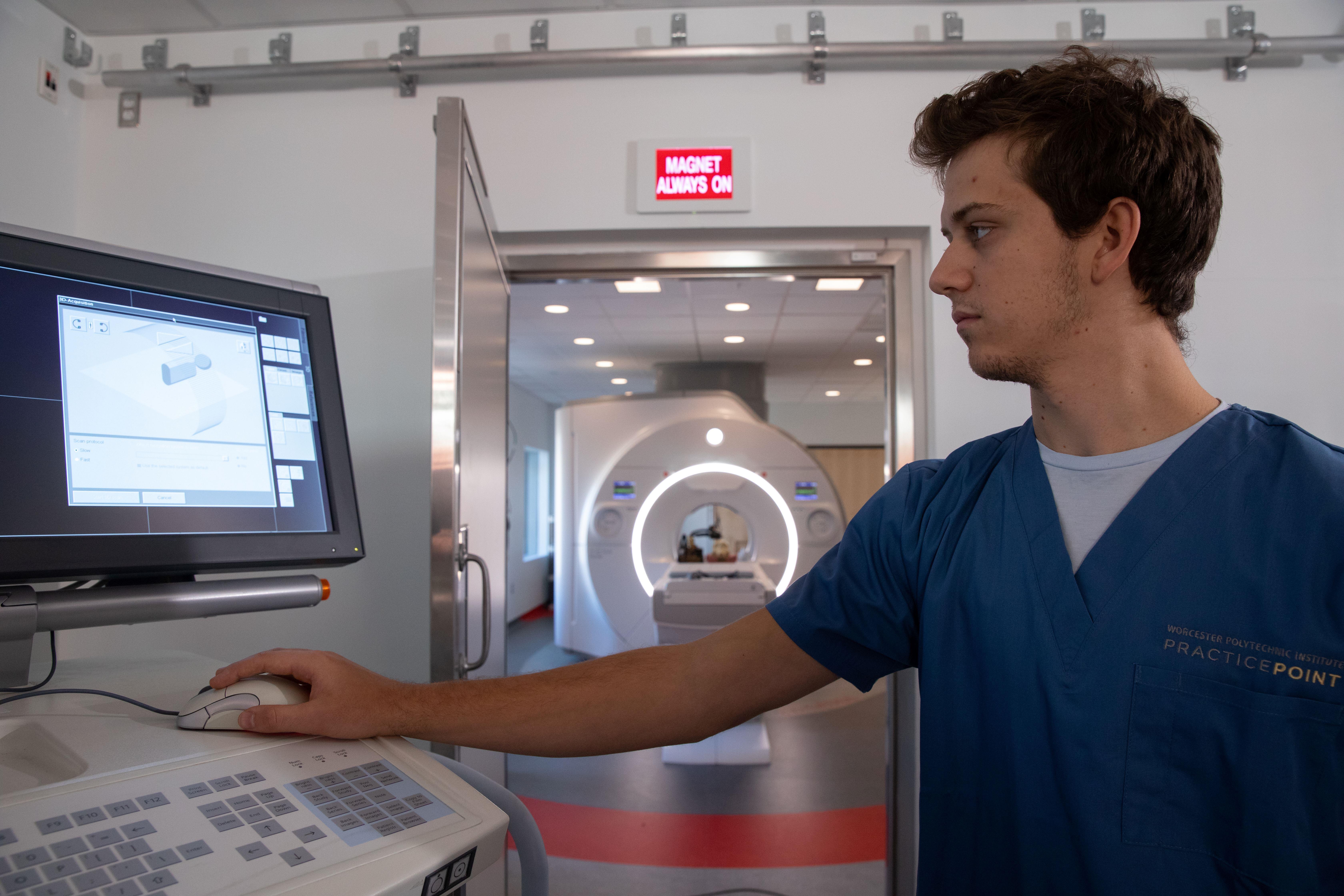 A student wearing scrubs working at a computer. In the background, an MRI machine is visible.