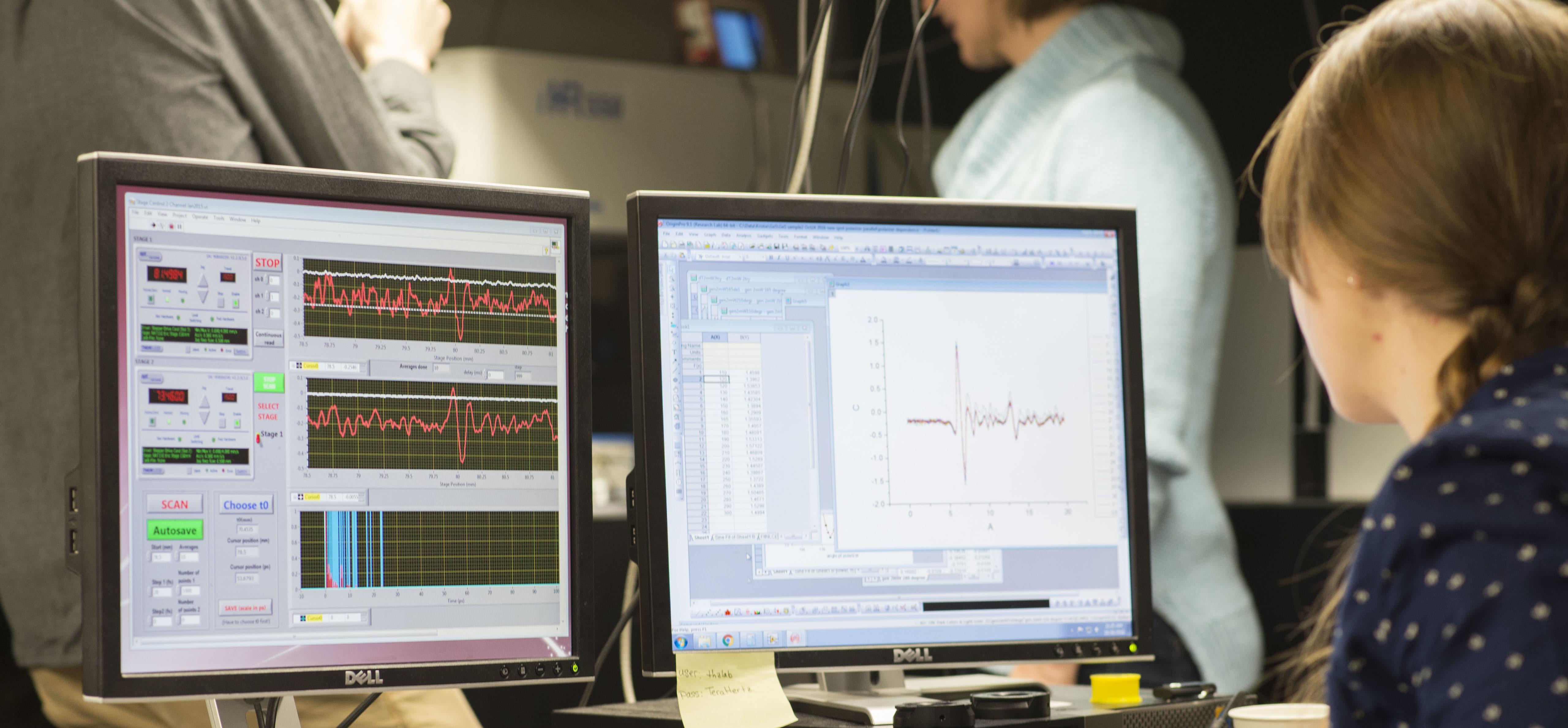 Woman reviewing data on two computer monitors with people in the background